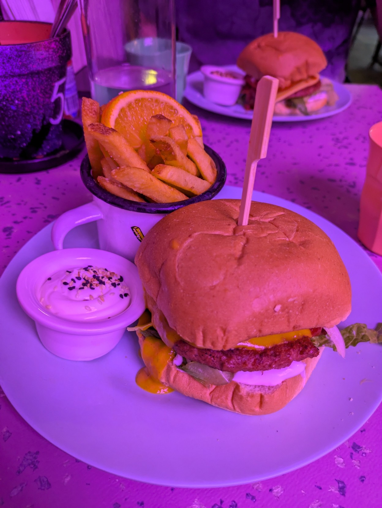 A vegan burger, fries with a lemon slice in a cup and a little cup of vegan mayo. The lighting of the photo is very purple.