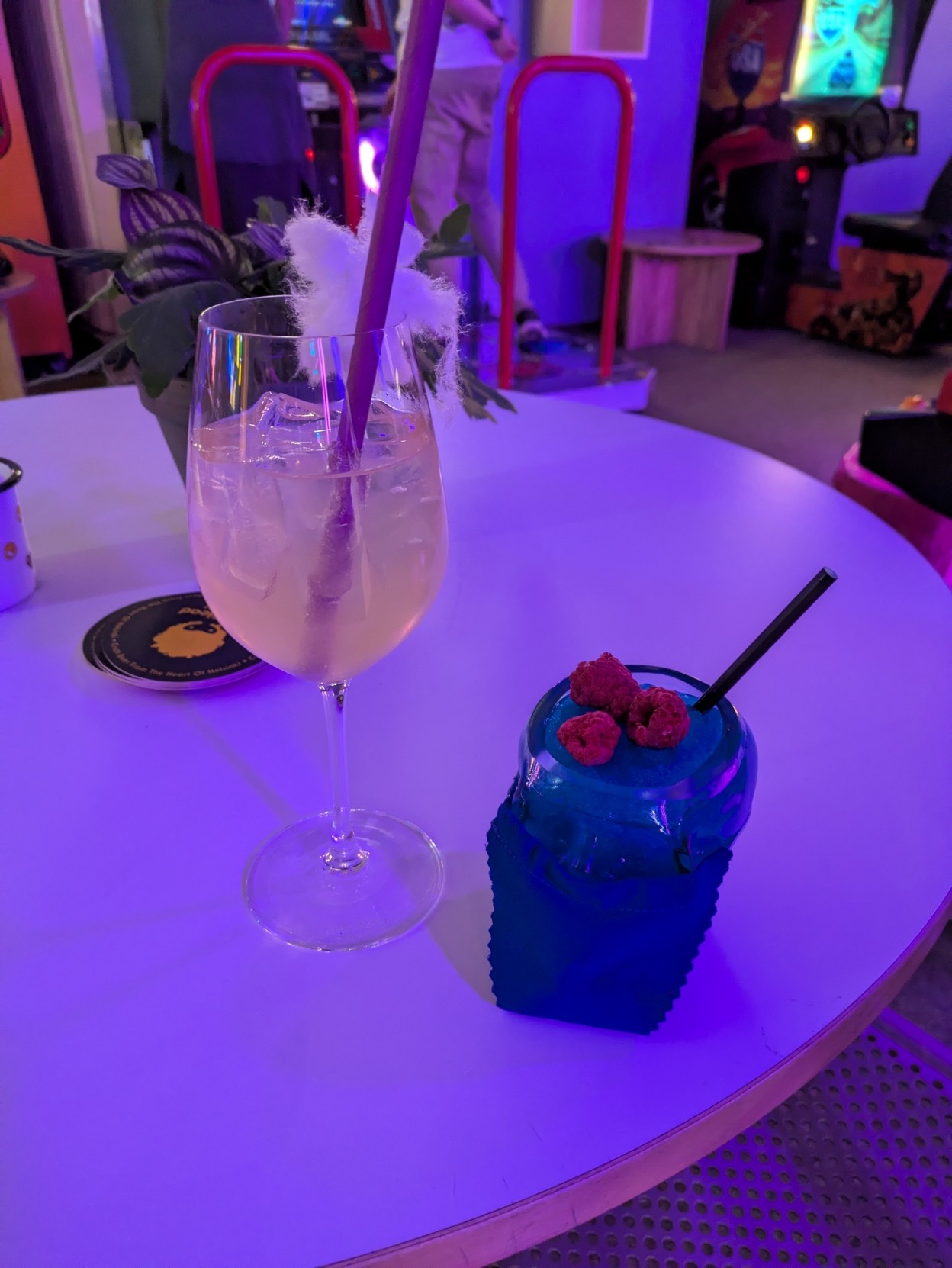Two mocktails on a table. Blue drink with raspberries is in a skull shaped glass and the other drink is light yellow color in a wine glass, with a bit of cotton candy on the edge of the glass.