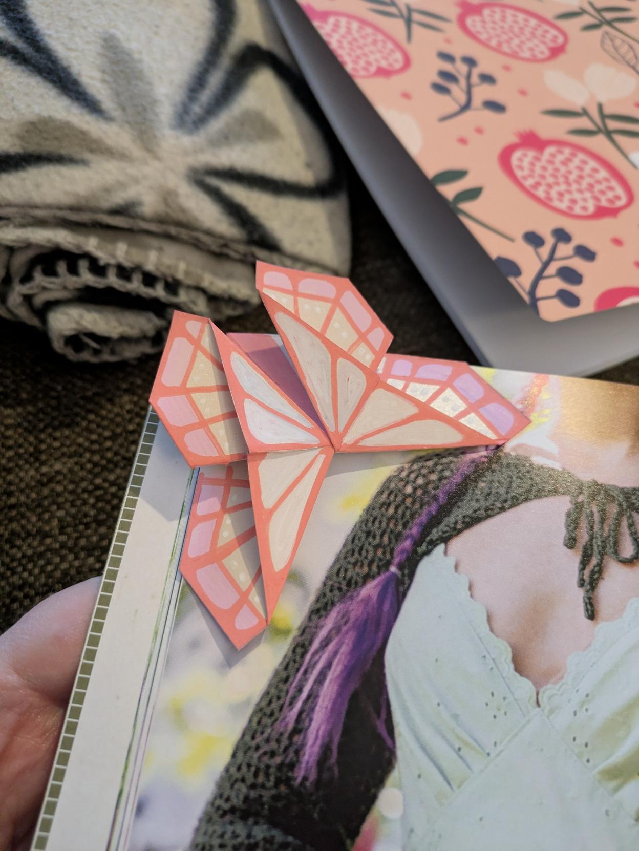 Butterfly shaped origami bookmark that's placed on the corner of the page. The butterfly is pale cream/white with pastel pink and coral colored details to make the wings look more butterfly-like
